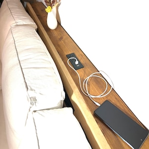 May include: A wooden sofa armrest with a built-in power outlet and USB port. A white charging cable is connected to a black smartphone. A white vase with dried flowers sits on the armrest.