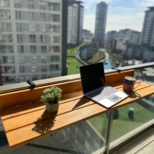 May include: A wooden balcony table with a laptop, a mug with an American flag design, and a small potted plant. The table is attached to a metal railing, with a city view in the background.