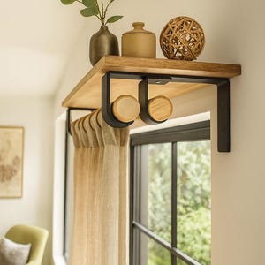 Op de afbeelding: Een houten plank met zwarte metalen beugels, met decoratieve items zoals een vaas, een kleine pot en een geweven bal. Daaronder zijn twee houten gordijnroedehouders zichtbaar, met beige gordijnen.