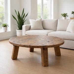 May include: A round coffee table constructed from weathered brown wood with a visible grain and texture. The table features a thick, circular tabletop supported by four sturdy, block-style legs, showcasing a natural, rustic appearance.