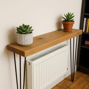 May include: A wooden console table with black hairpin legs. The tabletop is made of light-colored wood and supports two potted succulents. A white radiator is positioned below the table, and a bookshelf is visible in the background.
