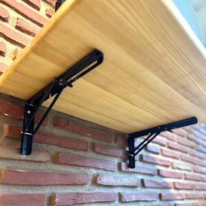 May include: A light-coloured wooden shelf supported by two black metal folding brackets. The brackets feature a triangular support arm and a horizontal mounting plate attached to the underside of the shelf, allowing the shelf to fold against the wall.