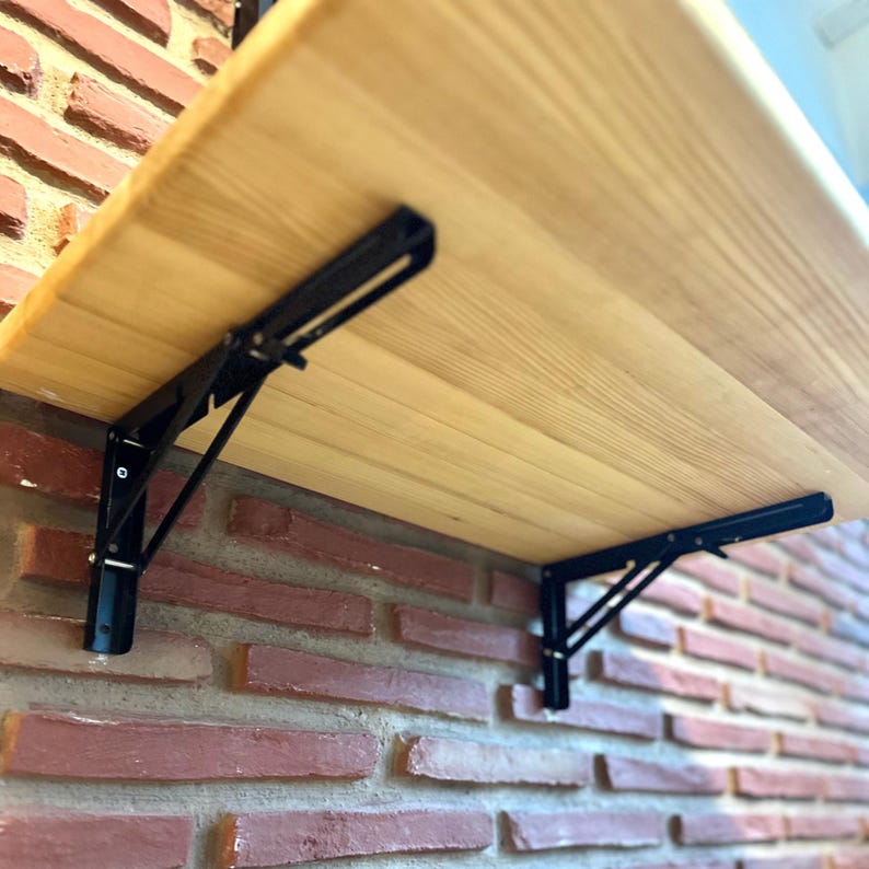 May include: A light-coloured wooden shelf supported by two black metal folding brackets. The brackets feature a triangular support arm and a horizontal mounting plate attached to the underside of the shelf, allowing the shelf to fold against the wall.