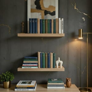 May include: A contemporary interior with two wooden shelves displaying a collection of books in shades of blue, green, and beige. A small plant, a white vase, and a wooden owl ornament are visible. A gold floor lamp and artwork are also present.