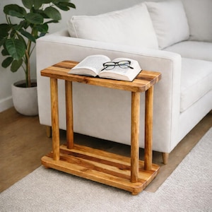 Op de afbeelding: Een rechthoekige houten bijzettafel met een natuurlijke houtnerfafwerking, voorzien van vier cilindrische poten en een onderste plank. Een open boek met witte pagina's en zwarte tekst ligt op het blad, met een bril met zwart montuur erbovenop.