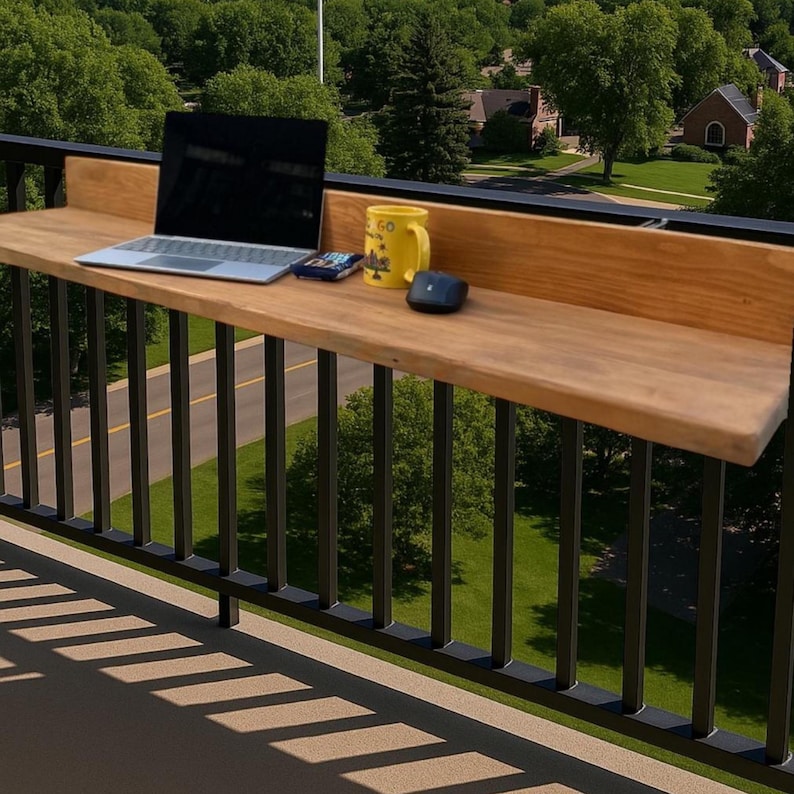 May include: A wooden balcony table with a laptop, a yellow mug, and a computer mouse on top. The table is attached to a black metal railing. The railing is in front of a green grassy area with trees and buildings in the background.