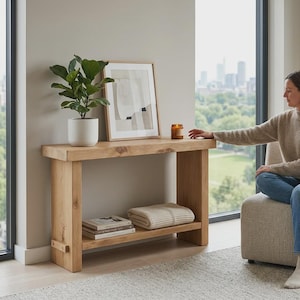 May include: A light-colored wooden console table with a lower shelf. A framed abstract art print, a potted plant, and a candle sit on top. A folded blanket and books are on the shelf. A person is sitting nearby.