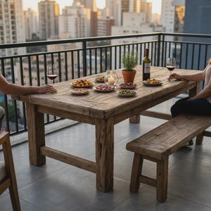 May include: A rectangular rustic wooden dining table with thick legs and a matching wooden bench. The table is set with several small plates of appetizers, two wine glasses, a bottle of red wine, and a small glass candle holder with a lit candle.