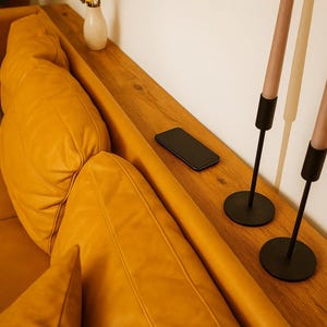 Op de afbeelding: Een close-up van een mosterdkleurige leren bank met een houten plank. Een zwarte smartphone ligt op de plank, samen met twee zwarte kandelaars met roze kaarsen. Een kleine witte vaas met een gouden accent is ook zichtbaar.