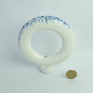 May include: White ceramic ring with blue speckles, standing on a small base. A coin is placed next to the ring for scale.