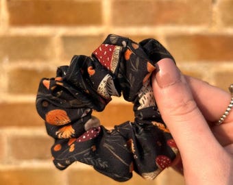 May include: Black scrunchie with a repeating pattern of mushrooms in red, orange, and white. The scrunchie is made of a soft fabric and is designed to hold hair in place. The background is a brick wall.