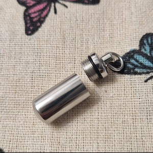 Puede incluir: Un colgante de metal plateado con un contenedor cilíndrico que se abre y cierra. El colgante tiene un bucle para sujetarlo a una cadena.