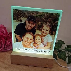 May include: A light-up photo frame featuring a family photo of four people. The frame is made of glass and has a wooden base. The photo shows a family of four in a natural setting. The text "Together we make a family!" is displayed on the frame.