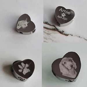 May include: Four silver heart-shaped cremation urns with engraved images and text. The urns are designed to hold ashes of a pet. One urn has a paw print, one has a dog's face, one has a cat's face, and one has the text "Arnie April 2021" with a paw print.