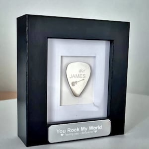 Puede incluir: Una caja de sombra enmarcada en negro con una púa de guitarra plateada en su interior. La púa tiene el nombre "JAMES" grabado en ella. La parte inferior de la caja tiene una placa plateada con el texto "You Rock My World "loving you - Scorpions" "