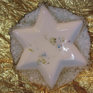 May include: A white, star-shaped dish with a decorative, ornate edge. The dish has a glossy finish and features delicate floral accents in shades of blue and green. The edge is detailed with a repeating pattern and small, gold-colored beads.