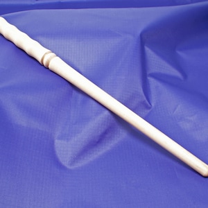 May include: A plain wooden wand with a light brown colour and a slightly darker brown band near the handle. The wand is lying on a blue fabric background.