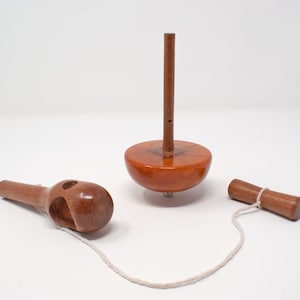 May include: A wooden spinning top toy with a brown body and a long, vertical brown peg. The toy is accompanied by a wooden handle and a pull string. The top is made of polished wood.