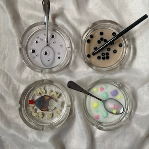 Puede incluir: Cuatro ceniceros de vidrio transparente con diferentes diseños. Un cenicero tiene un fondo blanco con ojos negros, otro tiene un fondo marrón con perlas de boba negras, otro tiene un fondo blanco con corazones de caramelo de colores y el último tiene un fondo blanco con un pez de dibujos animados y fideos.