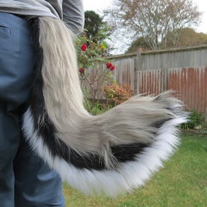 May include: A fluffy, gray, black, and white faux fur tail with a white tip. The tail is attached to a person's waist.