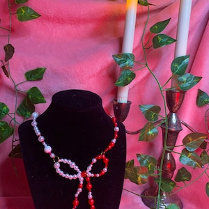 May include: A red and white pearl necklace with a bow in the center. The necklace is displayed on a black mannequin against a pink background with green vines and candles.