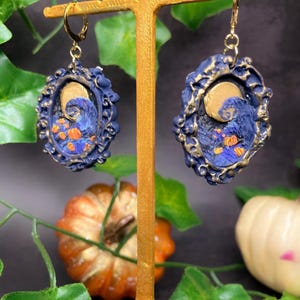 Peut inclure: Une paire de boucles d'oreilles sur le thème d'Halloween, de couleur bleue et dorée, représentant un croissant de lune et une silhouette de Jack Skellington du film L'Étrange Noël de monsieur Jack. Les boucles d'oreilles sont en pâte polymère et ont une finition dorée.