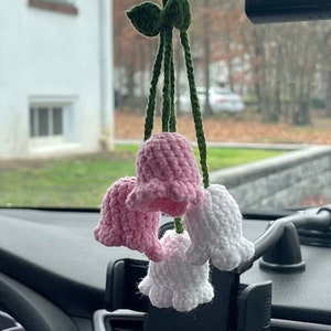 Op de afbeelding: Een autoluchtverfrisser met een tros gebreide roze en witte klokvormige bloemen die aan een groen touwtje hangen.