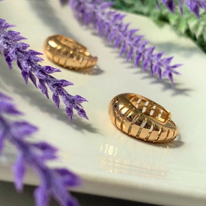May include: A pair of gold hoop earrings with a ridged design. The earrings are on a white surface with purple flowers.
