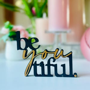 Könnte beinhalten: Ein schwarzes und goldenes Holzschild mit der Aufschrift "be you tiful". Das Wort "be" ist fett, "you" ist kursiv und "tiful" ist in Blockschrift geschrieben. Das Schild steht auf einer weißen Oberfläche. Weitere Objekte sind im Hintergrund.