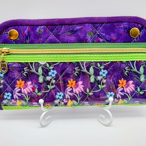 May include: A quilted purple wallet with a floral design, featuring a gold zipper and gold snap closures. The wallet has a green trim and a soft, velvety purple border. The floral pattern includes pink, orange, and blue flowers.