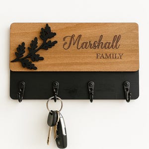 Könnte beinhalten: Schlüsselhalter aus Holz mit dem eingravierten Namen "Marshall" und dem Wort "FAMILY". Er hat eine schwarze Basis mit vier Haken und einem schwarzen Blattdesign. Schlüssel hängen an einem Haken.