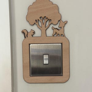May include: A wooden light switch cover with a nature theme. The cover features a tree, squirrel, and deer cut-out design. The switch plate is a brushed metal finish, and the frame is made of light-colored wood.