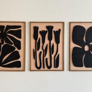 May include: Three wooden wall art pieces with black floral designs. The first piece features a large flower with multiple petals, the second piece features three stylized flowers with stems and leaves, and the third piece features a large flower with five petals.
