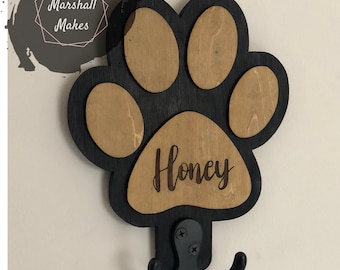 May include: A wooden paw print-shaped wall hook with the name "Honey" engraved on it. The paw print is black with natural wood-colored pads. A black metal hook is attached below. The text "Marshall Makes" is in a pink and white design.