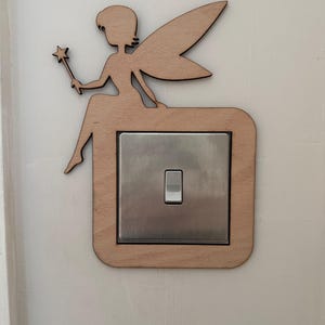 May include: A wooden fairy light switch cover. The fairy is holding a star-shaped wand. The light switch cover is square with rounded corners and a silver switch. The fairy is sitting on top of the light switch cover.
