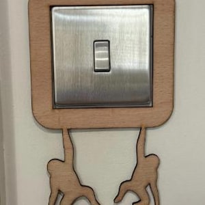 May include: A light wood-toned decorative light switch cover with a stainless steel switch. Two monkey silhouettes are hanging from the bottom of the cover. The cover is square with rounded corners.