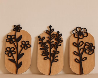 May include: Three wooden wall art pieces featuring black floral designs. Each piece has a light brown, oval-shaped background. The floral designs include flowers and leaves, creating a natural, decorative theme.