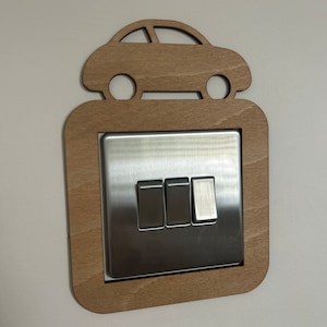 May include: A wooden light switch cover with a cutout of a car on top. The switch cover is a light brown color and the car is a darker brown color. The switch cover has three light switches.