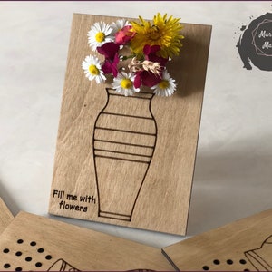 Może przedstawiać: Drewniane rzemiosło z konturem wazonu i tekstem "Fill me with flowers". Wazon jest ozdobiony kwiatami, w tym białymi stokrotkami, żółtymi kwiatami i czerwonymi kwiatami.