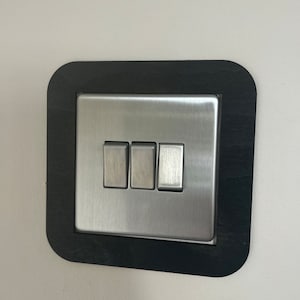 May include: A brushed stainless steel light switch with three toggle buttons, framed in a black square.