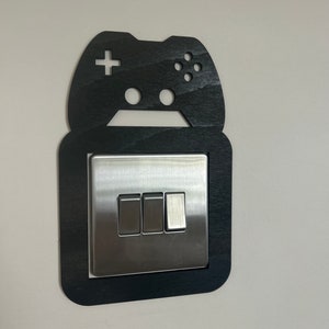 May include: A black wooden light switch surround featuring a cutout in the shape of a video game controller. The decorative frame is mounted around a rectangular metallic triple light switch plate with three vertical toggle switches.