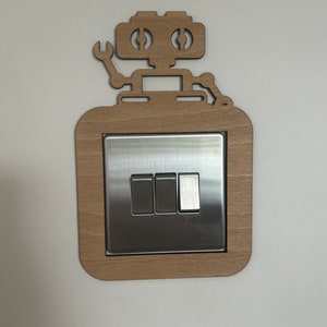 May include: A wooden robot-shaped light switch cover with a silver switch plate. The robot has a wrench arm and a square body. The cover is made of light-colored wood and is mounted on a white wall.