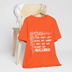 May include: Bright orange t-shirt featuring the text "And winter gave spring and summer a miss and went straight into Autumn" in white. A white cap is on a wooden chair.