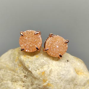 Pode incluir: Um par de brincos redondos com uma superfície brilhante de cor pêssego. Os brincos são fixados em um metal de ouro rosa com quatro garras que prendem a pedra. Os brincos são exibidos em uma superfície clara e texturizada.