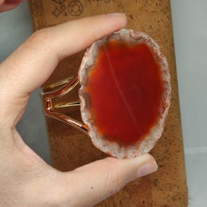 May include: A cuff bracelet featuring a large, polished, translucent orange-red agate slice with a natural, rough, light-colored crystalline edge. The stone is securely mounted onto a shiny, polished gold-tone metal band with a split design.