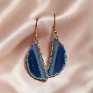 Può includere: Un paio di orecchini pendenti con fette di agata blu semicircolari lucide con venature bianche naturali. Ogni pietra è incastonata in un bordo di metallo color oro testurizzato e pende da un semplice gancio color oro per lobi forati.