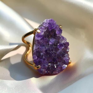 Pode incluir: Um anel de tom dourado com uma grande pedra de ametista em forma de lágrima. A superfície cristalina roxa profunda da gema é mantida por uma cravação de metal polido, fixada a uma banda dourada lisa, curva e com design aberto.