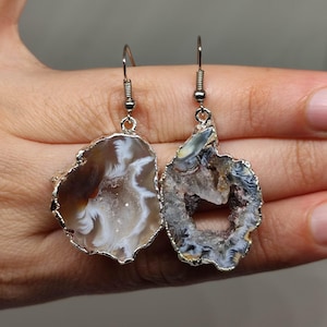 May include: A pair of natural stone earrings with silver-coloured metal trim. The earrings have a unique, organic shape with brown, white, and grey patterns. The earrings are suspended from silver-coloured ear wires.