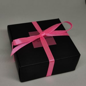 Puede incluir: Una caja de regalo negra con una cinta rosa atada en un lazo. La caja tiene una etiqueta que dice "Love Jewels".