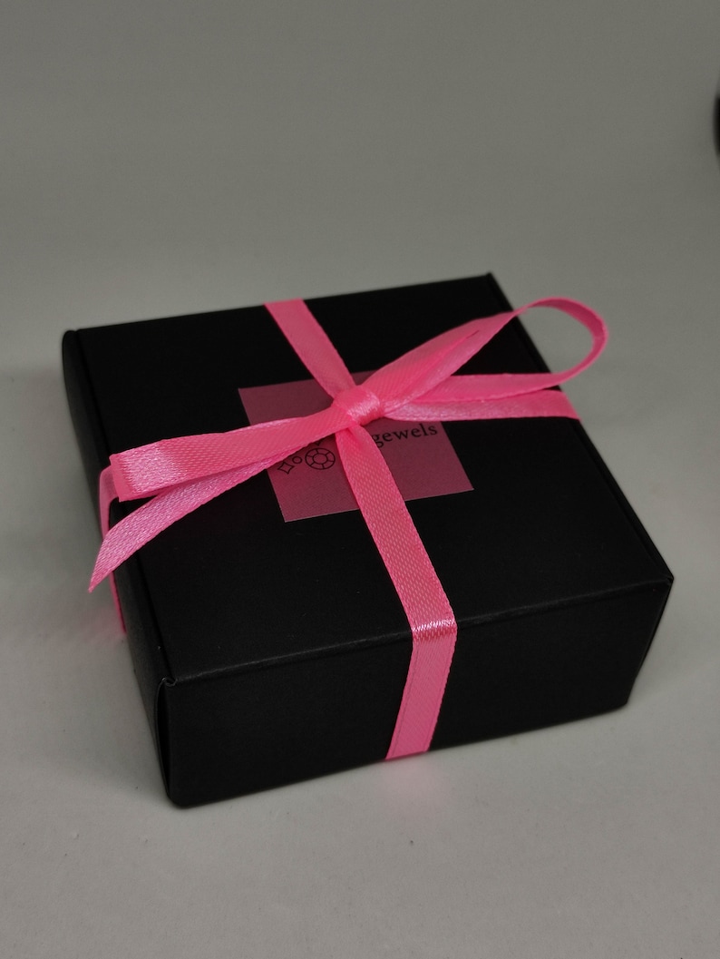 Puede incluir: Una caja de regalo negra con una cinta rosa atada en un lazo. La caja tiene una etiqueta que dice "Love Jewels".